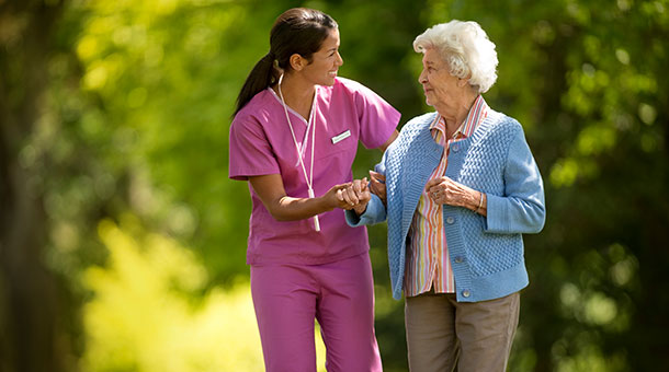 A nurse walks with an older woman through a park.