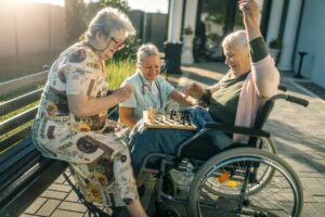 Two friends, one in a wheelchair, play chess alongside a home health aide