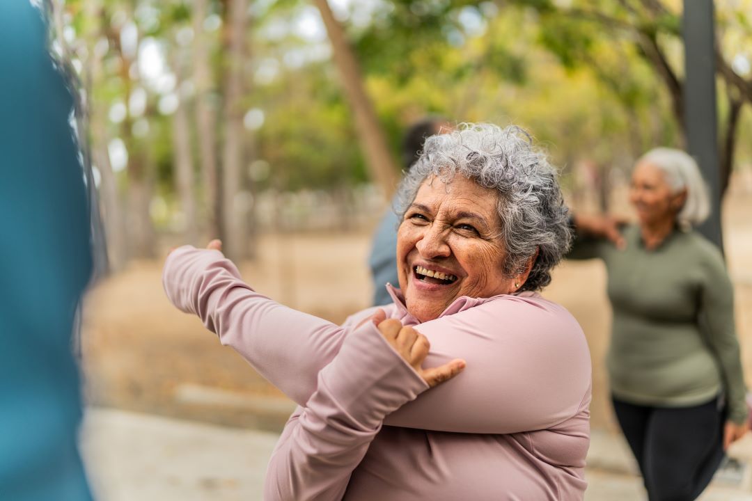 An older woman smiles and stretches outdoors as she prepares to have fun with friends.